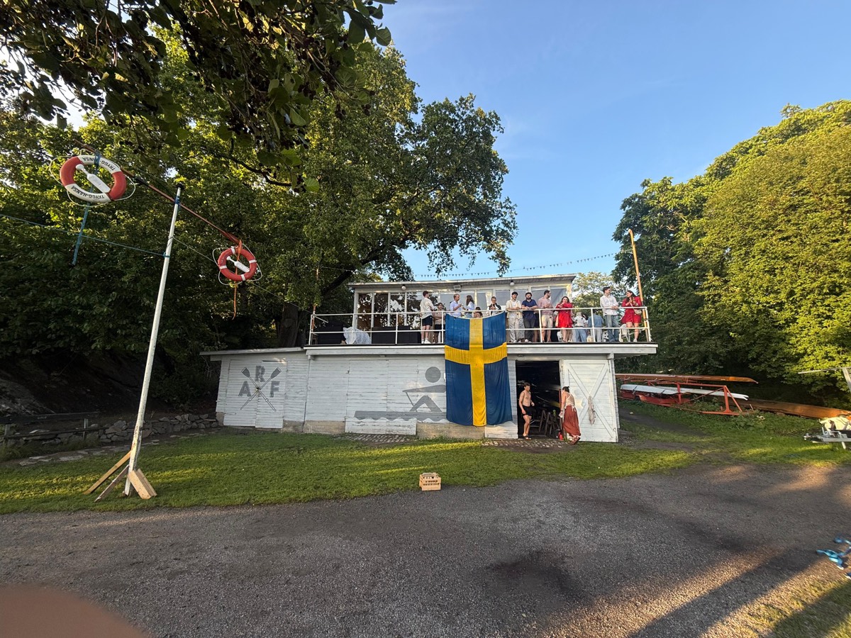 A white boathouse at dusk with a large Swedish flag over the doors and people on the balcony