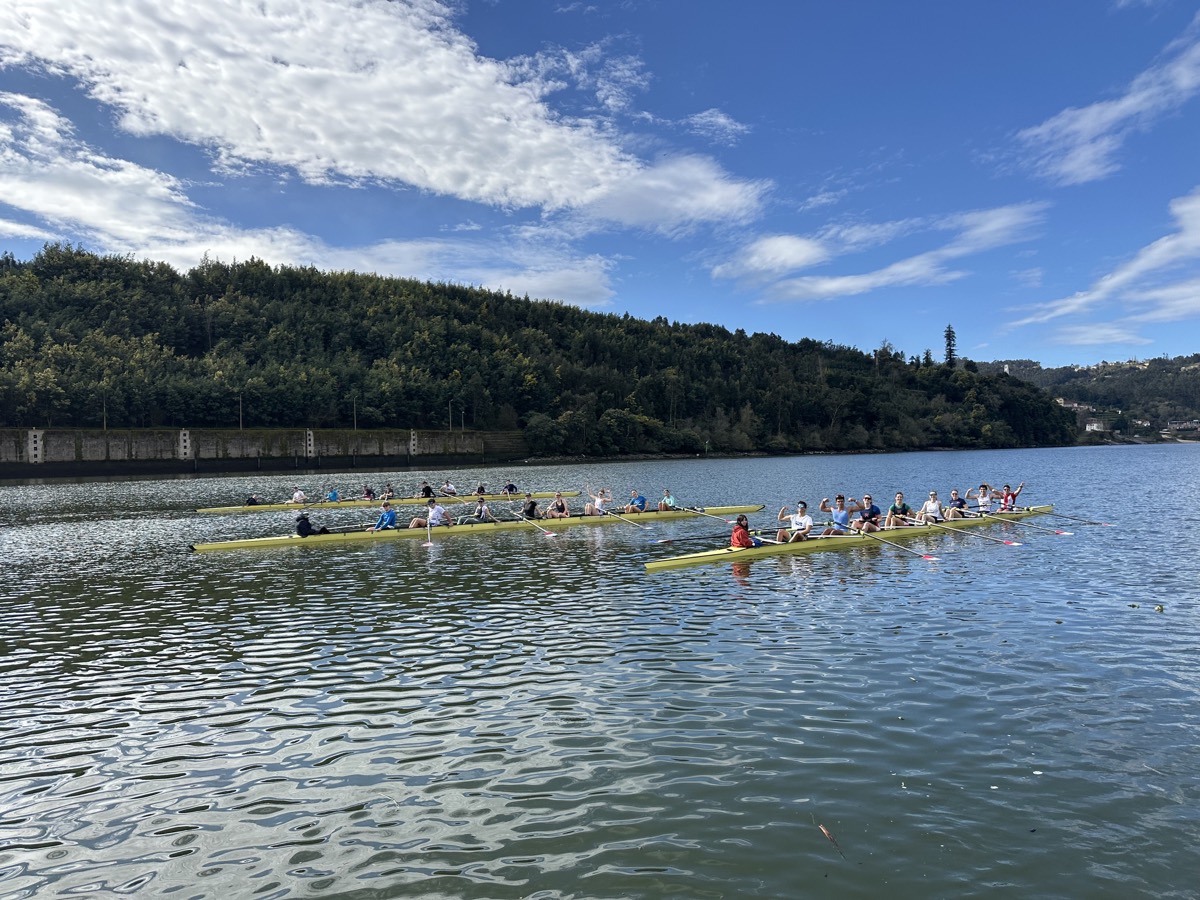 Several crews in eights and quads gathering on a sunlit river below forested hills