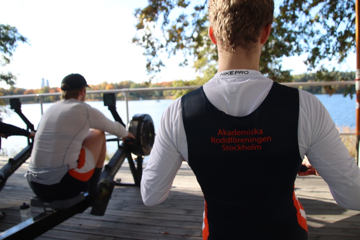 Two rowers on ergometers on a dock overlooking the water, club jersey reading Akademiska Roddföreningen Stockholm