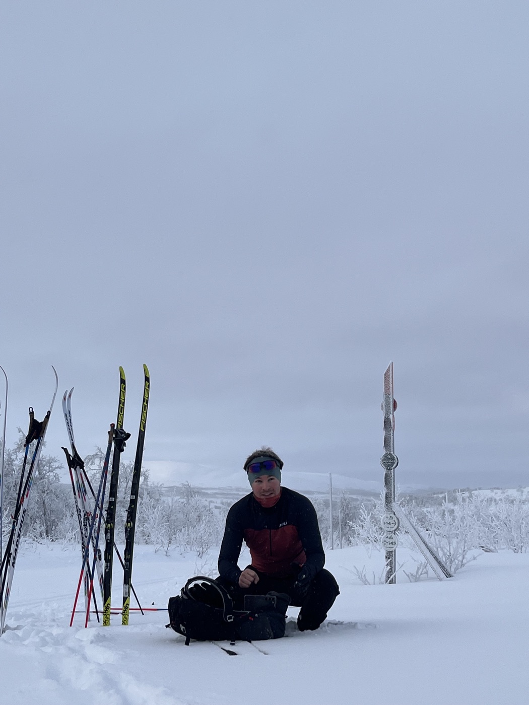 A ski tourer crouched over a backpack in a vast snowy fjäll plain, skis planted upright